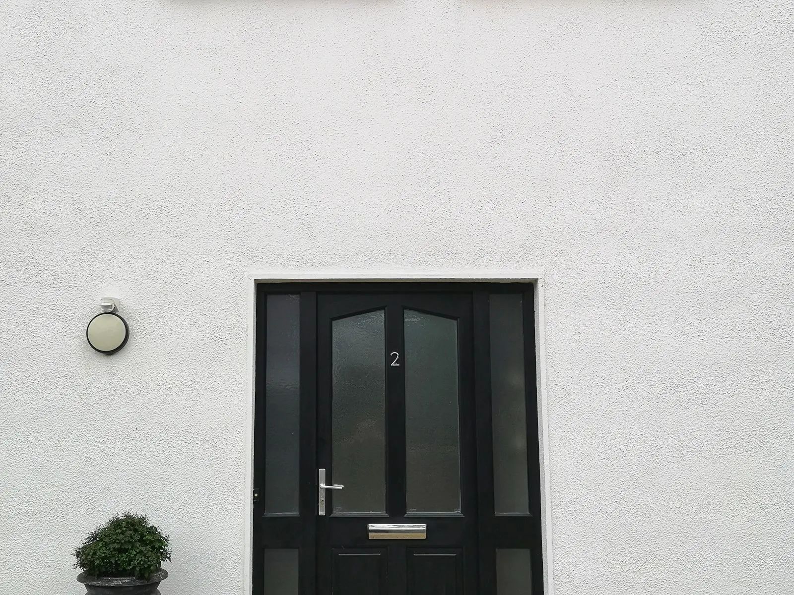 Beyaz sıvalı villa cephesinde yan camlı siyah giriş kapısı; kanadın üstünde 2 numarası, yanında duvar apliği ve saksı.