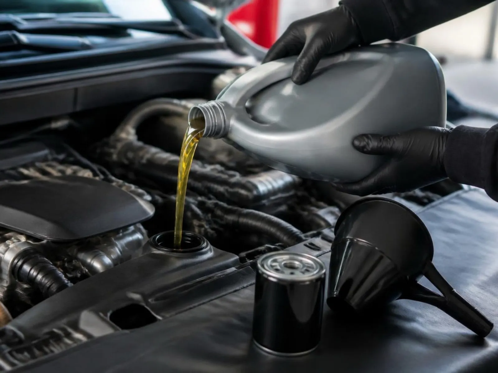 Açık motor kaputunun altında yağ filtresini değiştiren eldivenli bir el; yanında yeni filtre ve yağ bidonu duruyor.